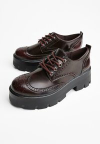 Un paio di scarpe brogue in pelle marrone scuro con suole nere spesse e lacci marroni coordinati su una superficie bianca.