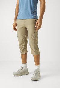 Shorts cargo beige avec plusieurs poches, coupe droite, en tissu léger. Associés à des chaussures de sport grises et des chaussettes blanches. Chemise bleue en haut.