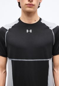 Νεαρός ενήλικας με κοντά μαλλιά που φοράει αθλητικό μπλουζάκι Under Armour σε μαύρο και γκρι χρώμα, εμφανίζεται από τους ώμους μέχρι το μέσο του κορμού σε λευκό φόντο.