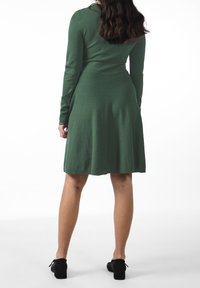 Robe verte à manches longues en matière douce, ajustée en haut et évasée à la taille, avec des accents en dentelle aux manches. Des chaussures noires complètent le look.