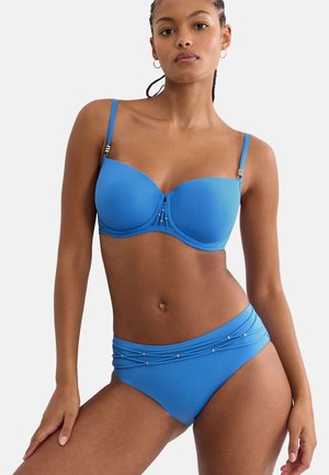 Frau mit geflochtenem Haar, die einen blauen Bikini mit goldenen Perlendetails am Ober- und Unterteil trägt, vor einem schlichten Hintergrund stehend.