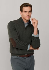 Donkergroene trui met een ritskraag, geribbelde manchetten en bruine elleboogpatches, gelaagd over een lichtblauw en wit gestreept shirt.