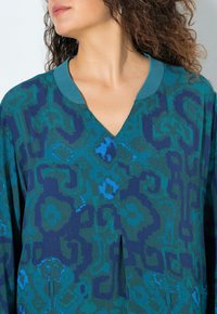 Blusa de manga larga en tejido azul-verde texturizado con escote en V y un cuello en contraste. El patrón incluye formas irregulares y acentos.