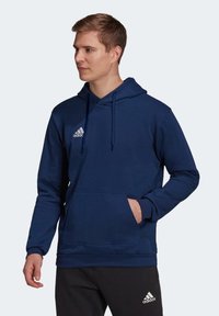Mørkeblå hettegenser med lommer, justerbar hette og hvit logo på brystet; laget av mykt stoff med lange ermer.