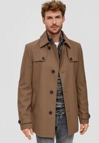 Brauner, strukturierter Mantel mit hohem Kragen, Knopfverschluss und zwei seitlichen Taschen. Getragen über einem gemusterten grauen Pullover und Jeans.