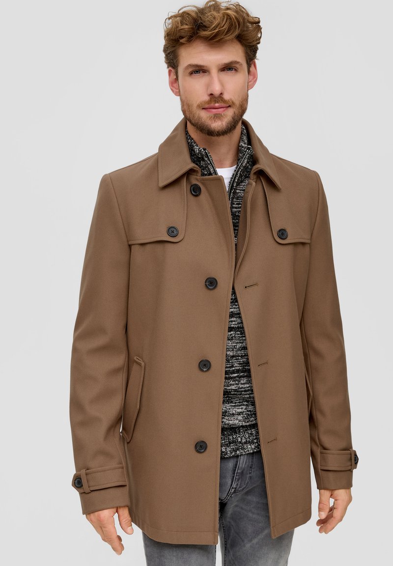 Brauner, strukturierter Mantel mit hohem Kragen, Knopfverschluss und zwei seitlichen Taschen. Getragen über einem gemusterten grauen Pullover und Jeans.