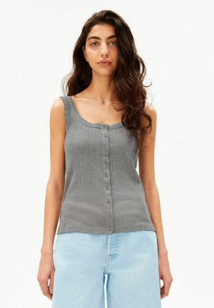 Femme aux longs cheveux ondulés portant un haut gris sans manches boutonné et un jean bleu clair, debout devant un fond blanc uni.