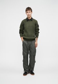 Oliivinvihreä ribbipintainen neule mustalla kauluksella, yhdistettynä vaaleanharmaisiin cargo-housuihin, joissa on useita taskuja ja vetskareita, mustien lenkkarien kanssa.