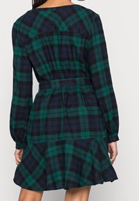 Robe à carreaux verts et bleu marine avec manches longues, ceinture à la taille, jupe évasée et poignets boutonnés. Tissu doux avec finition texturée.