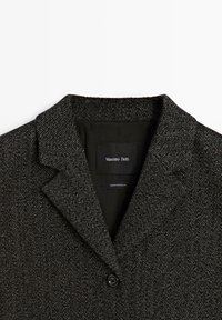 Blazer gris foncé à motif en chevrons, avec un col à revers cranté et un bouton noir unique à l'avant.