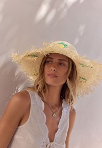 Femme portant un chapeau en paille à larges bords avec des citrons brodés et un haut sans manches blanc avec des bords festonnés, tournée légèrement vers la gauche.