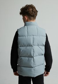 Svijetloplava bezrukavna puffer prsluk s kvadratnim uzorkom, visokim ovratnikom i glatkom teksturom, nosi se preko crne košulje s dugim rukavima.