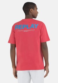 Punainen puuvillainen t-paita, jossa on suuri sininen "REPLAY" -logo selässä, pyöreä kaula-aukko ja lyhyet hihat. Ihanteellinen rentoon käyttöön.