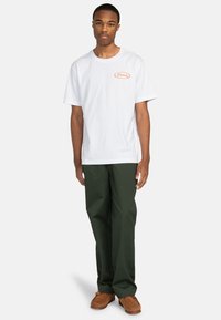 Hvid bomulds-T-shirt med et rødt ovalt logo på brystet, parret med mørkegrønne bukser og brune mokkasinstøvler. Modellen står med fronten vendt mod kameraet.