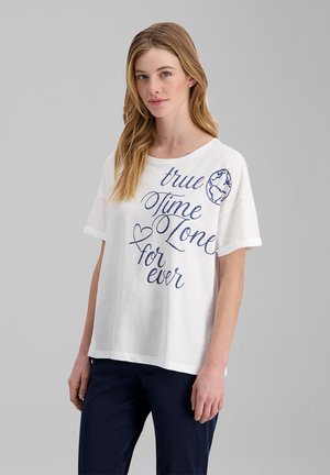 Young woman with long light brown hair wearing a white T-shirt with "true Time Zone for ever" text and a globe graphic, paired with dark pants.