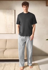 Czarny t-shirt z krótkim rękawem w połączeniu z niebieskimi i białymi pasiastego wzoru spodniami piżamowymi. Model stoi boso w neutralnym wnętrzu.