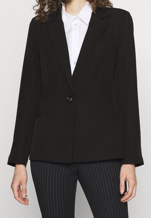 Femme portant un blazer noir cintré sur une chemise blanche à col et un pantalon noir à fines rayures, debout devant un fond uni.