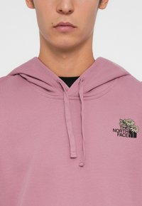 Lyserød hoodie lavet af blødt stof. Har en frontkangaroo-lomme, snoretræk til hætten og broderet logo med blomstermotiv på brystet.