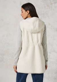 Fleece vest, crème kleur, met capuchon, gestructureerde stof, tailleband, gelaagd over een grijze long-sleeve shirt, blauwe spijkerbroek, gezien vanuit de rug.