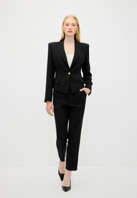 Černý na míru šitý oblek s elegantním sako a zlatým knoflíkem, doplněný rovnými kalhotami. Tkanina se zdá být hladká a lesklá.