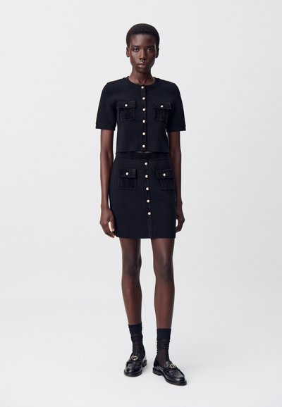 Femme portant un cardigan à manches courtes noir à boutons et une jupe assortie avec des poches avant, associée à des chaussettes noires et des mocassins.