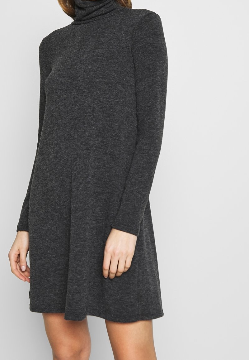 Femme portant une robe pull à col roulé gris foncé à manches longues, debout devant un fond clair uni.