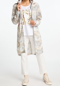 Długi, jasnoszary i beżowy płaszcz przeciwdeszczowy w kamuflażu z zamkiem z przodu, w połączeniu z białą koszulą z nadrukiem w kolorze złotym oraz białymi spodniami.