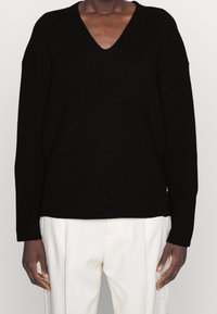 Svart stickad tröja med V-hals, lös passform och långa ärmar. Strukturerad tyg och sidoriginaler vid fållen. Kombinationen av vita byxor.