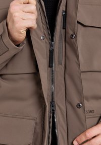 Brun isolerad jacka med blixtlåsförsedd fram, tryckknappar och texturerat tyg. Har flera fickor och kontrasterande hårdvarudetaljer.