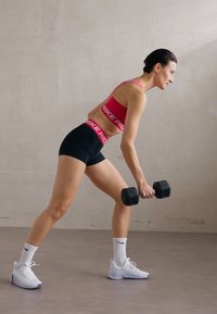 Rozā sporta krūšturis ar "NIKE PRO" marķējumu, melni šorti, turēšana sešstūra hanteli. Baltas sporta kurpes un zeķes. Neitrāls fons.