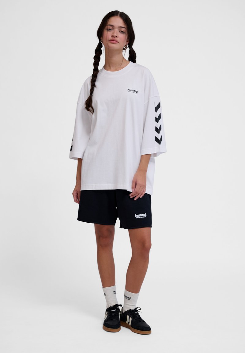 Bílý oversized tričko s černými chevronovými akcenty na rukávech, spárované s černými sportovními šortkami s logem. Černé tenisky a bílé ponožky.