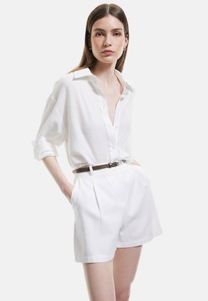 Valkoinen pellavapaita nappilista, käärittyjä hihnoja ja korkeavyötäröiset valkoiset shortsit, joissa on sartorial-pleatit. Ruskea vyö tuo kontrastia.