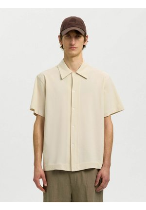 Jeune homme portant une chemise beige à manches courtes boutonnée, une casquette marron et un pantalon vert olive, debout devant un fond clair uni.
