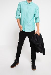 Minzgrünes Hemd mit Knopfleiste und Brusttasche, kombiniert mit schwarzen, schlanken Hosen. Schwarze Schuhe mit braunen Sohlen, hält eine schwarze Jacke.