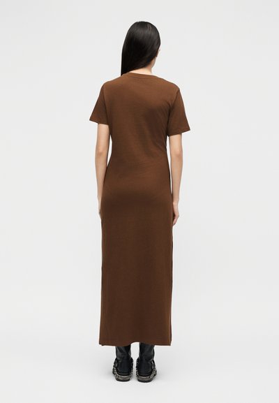 Γυναίκα με μακριά σκούρα μαλλιά, που φοράει ένα καφέ μάξι φόρεμα με κοντά μανίκια και μαύρες μπότες, στέκεται με την πλάτη γυρισμένη σε ένα απλό φωτεινό φόντο.