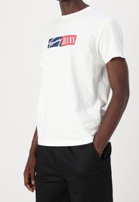 Biała bawełniana koszulka z nadrukiem graficznym z napisem "Tommy Jeans" w niebieskim i czerwonym kolorze. Klasyczny krój z okrągłym dekoltem i krótkimi rękawami.