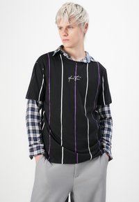 Joven con cabello corto rubio que lleva una camiseta de rayas negra sobre una camisa de cuadros azul de manga larga y pantalones gris claro, con las manos en los bolsillos.