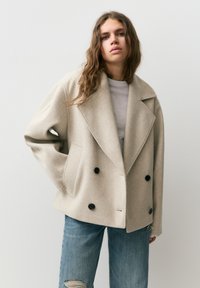 Młoda kobieta z długimi brązowymi włosami, ubrana w beżowy płaszcz dwurzędowy i podarte niebieskie dżinsy, stojąca na tle jednolitego jasnego tła.