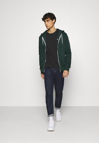 Grønn hettejakke med hvite snorer over en svart T-skjorte, kombinert med mørkeblå jeans og hvite joggesko. Har sidelommer.