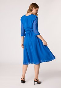 Blauwe chiffon jurk met een fitted taille, driekwart mouwen en een vloeiende rok. Achterkant beschikt over een sleutelgatsluiting. Gestyled met zwarte hakken.