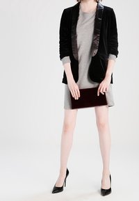 Černý sametový blejzr s lesklými saténovými klopami přes světle šedé šaty, doplněný vínově červeným psaníčkem a černými lodičkami na podpatku.