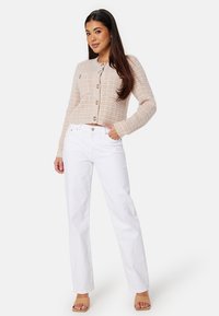 Beige textuur cardigan met knopen aan de voorkant, gecombineerd met hoge witte jeans. Model staat en toont beide kledingstukken en lichtbeige sandalen.