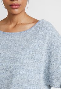Pull en tricot bleu clair avec un col asymétrique, coupe ample et motif texturé. Matériau doux avec des variations de couleur subtiles.