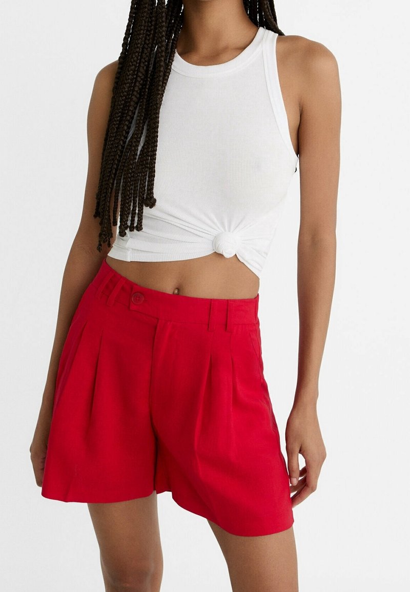 Femme portant un crop top blanc sans manches noué à la taille et un short plissé rouge taille haute sur un fond uni.