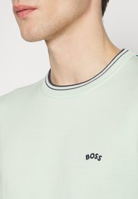 Gros plan d’un jeune homme portant un T-shirt vert clair avec un col rayé bleu marine et « BOSS » brodé sur la poitrine.