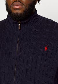 Mörkblå kabelstickad tröja med hög krage, med en gul dragkedja och en liten röd logotyp på bröstet. Strukturerad, ribbad design.