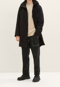 Black waterproof coat with a hood over a beige sweater, paired with dark jeans featuring white stitching and black lace-up boots.