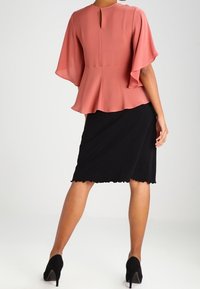 Persikka blusa, jossa on lepattelevat hihat, pyöreä selkä ja peplum-helma, yhdistettynä mustaan polvipituiseen hameeseen, jossa on aaltoileva reuna.