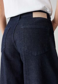 Jean en denim bleu foncé avec une coupe droite, comportant deux poches arrière et une étiquette de marque en cuir sur la taille.