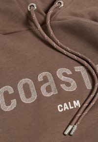Sweat à capuche en coton marron avec les mots « coast » et « calm » brodés en contraste, et cordons de serrage avec embouts métalliques argentés.